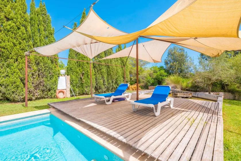 A swimming pool beside a wooden deck with lounge seating beneath shade sails.