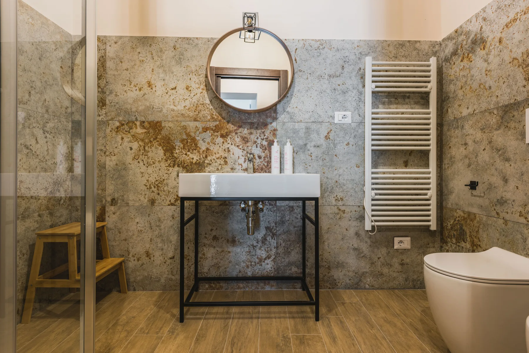 Un baño con lavabo suspendido, espejo redondo y toallero calefactable.