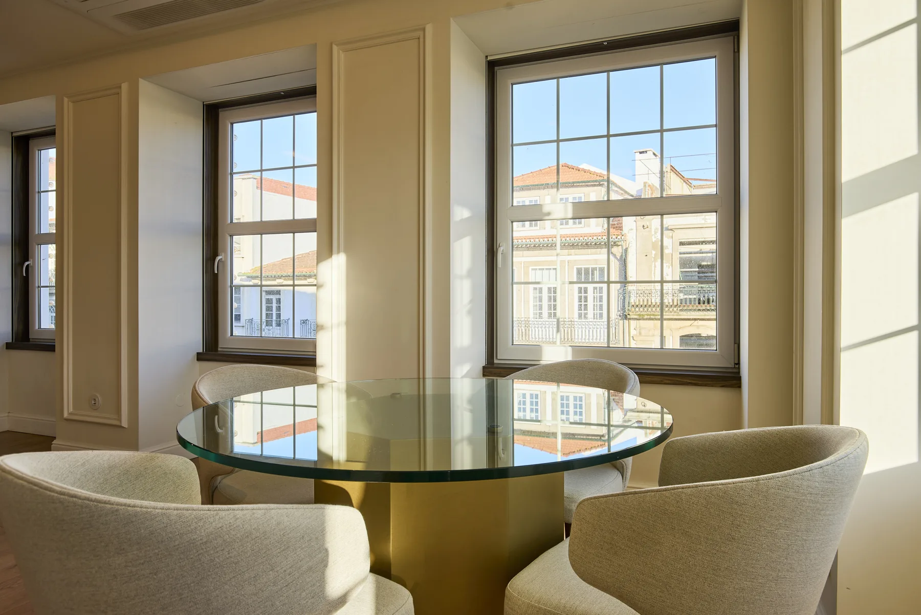 A dining area with a round glass table and upholstered seating beside large windows.