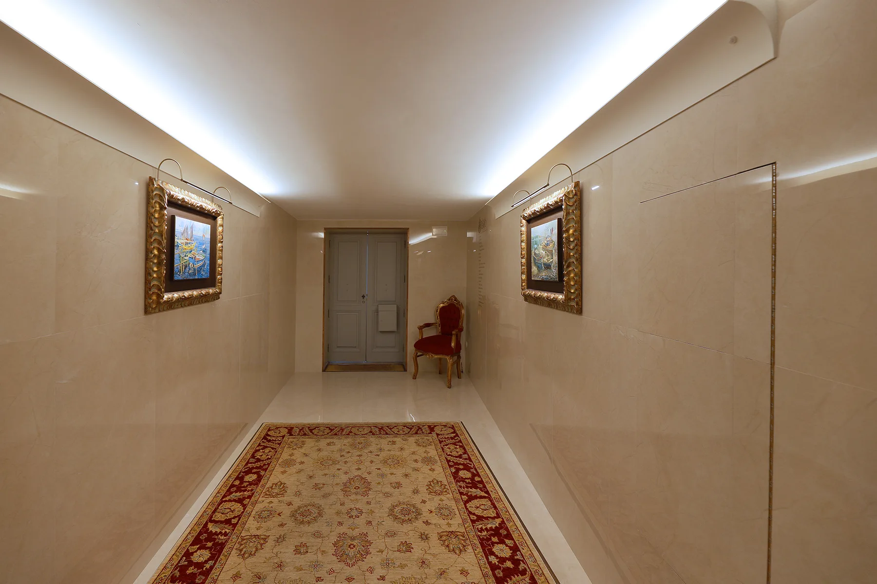 A narrow hallway with framed artwork, a patterned rug, and a chair by the far door.