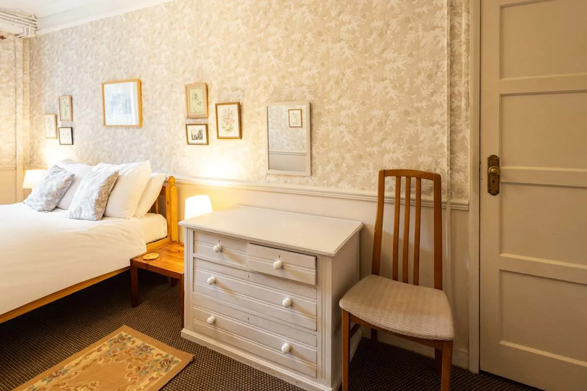 A bedroom with a bed, dresser, and chair against patterned wallpaper.