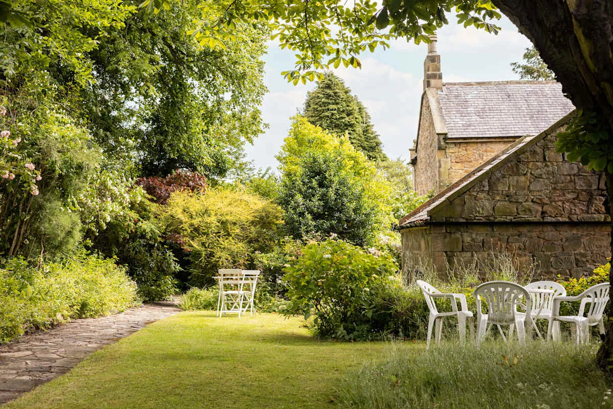 A garden seating area beside a stone building, surrounded by trees and greenery.