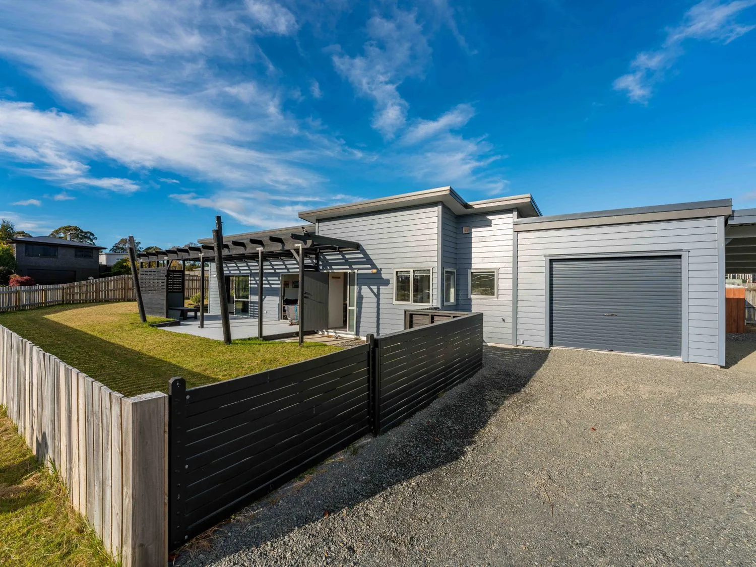 Una casa moderna con paredes exteriores grises, garaje, un jardín cercado y una pérgola bajo un cielo azul en Motuoapa Getaway Lake Taupo Holiday Home, en Motuoapa