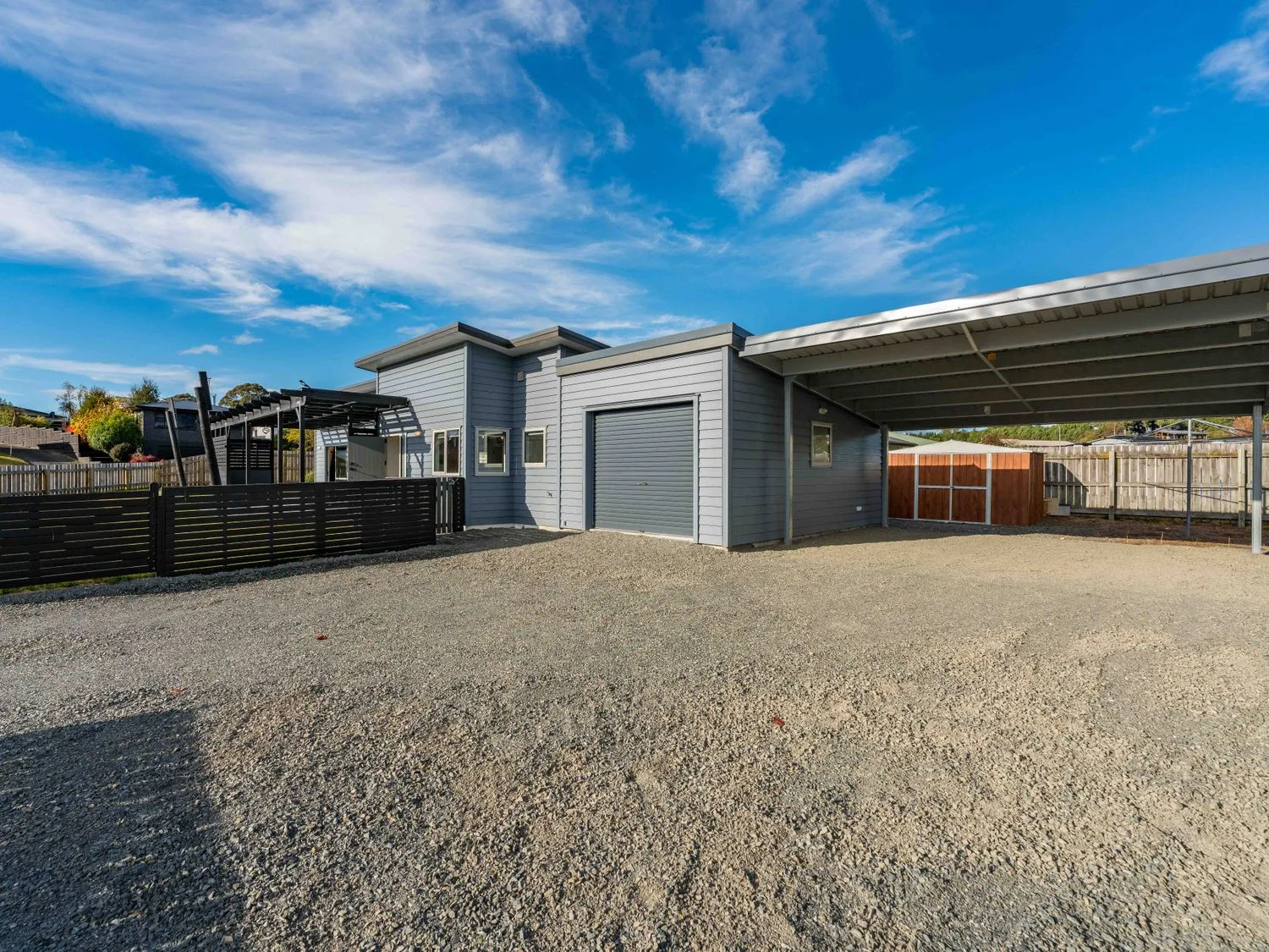 An outdoor area with a gravel driveway a carport a grey garage door a wooden fence and a shed at Motuoapa Getaway Lake Taupo Holiday Home Motuoapa