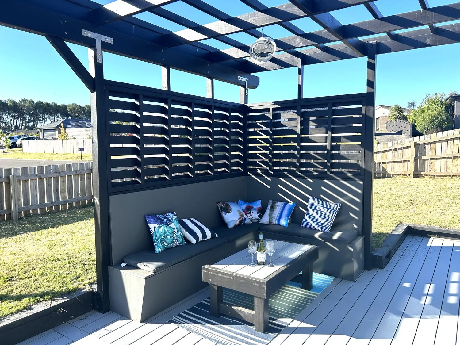 Una zona de estar al aire libre con un banco esquinero con cojines y una mesa con copas de vino y una botella bajo una pérgola en Motuoapa Getaway - Lake Taupo Holiday Home, en Motuoapa