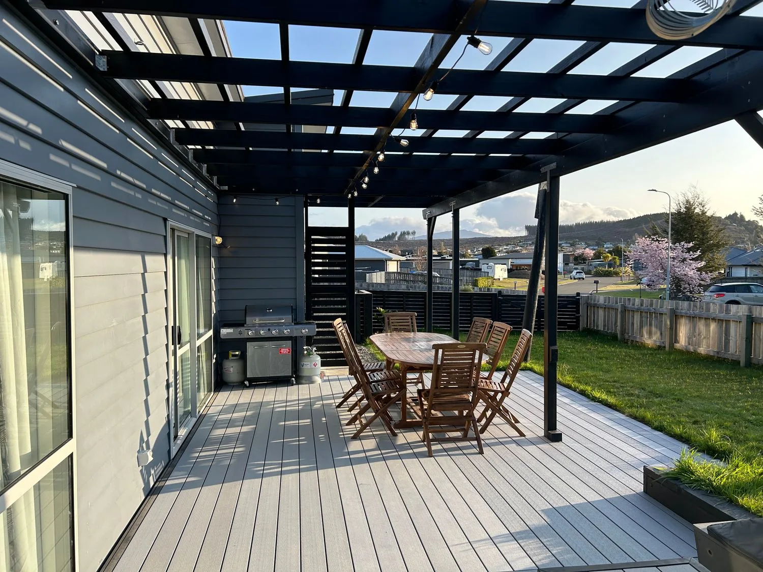 Un patio al aire libre con una mesa de comedor y sillas de madera bajo una pérgola con una guirnalda de luces y una parrilla en Motuoapa Getaway - Lake Taupo Holiday Home, en Motuoapa