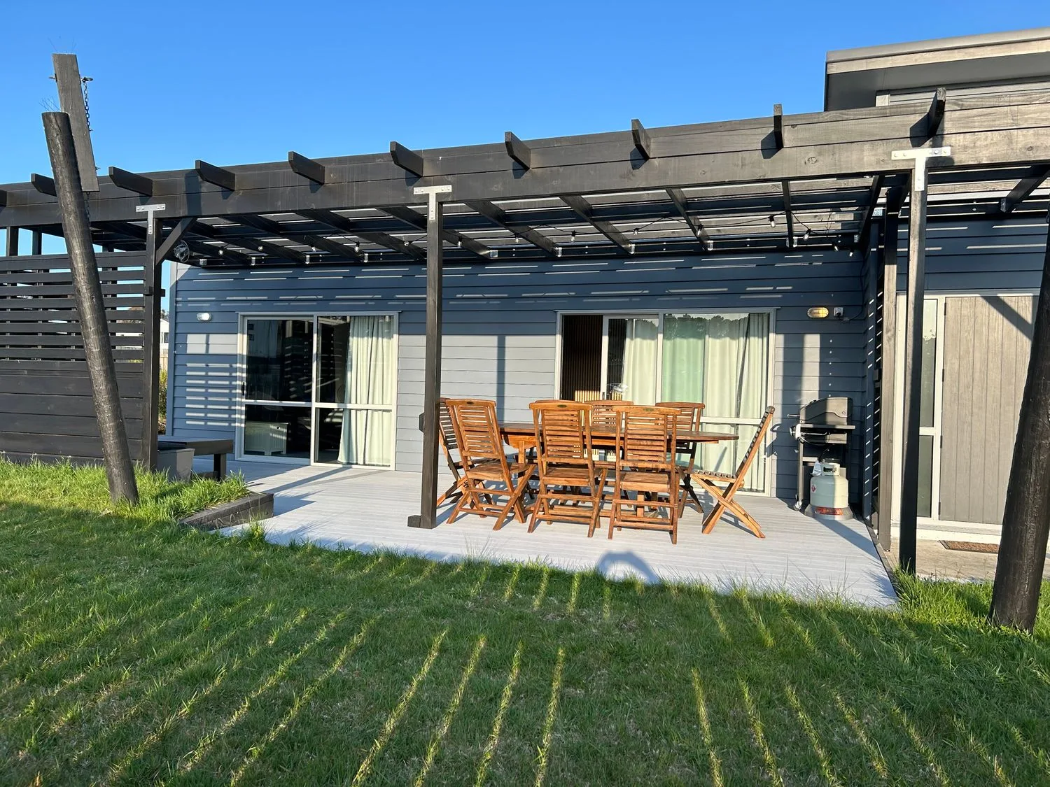 An outdoor patio with wooden seating sits beneath a pergola beside a modern home.