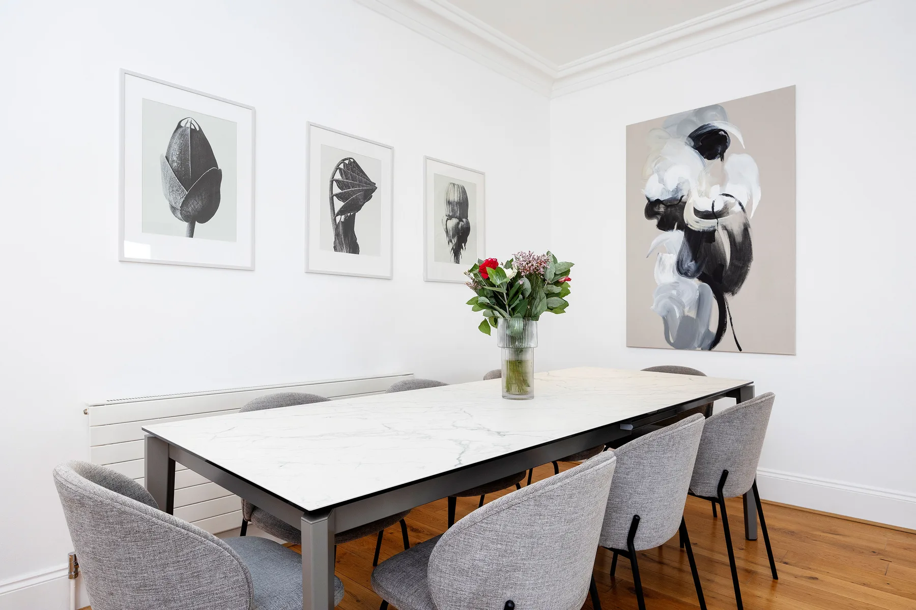Comedor con una mesa, asientos grises, cuadros enmarcados y flores.