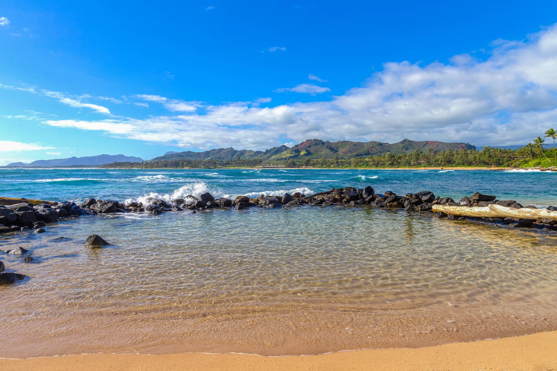 Una playa de arena se encuentra con una orilla poco profunda y tranquila, con rocas y colinas lejanas.