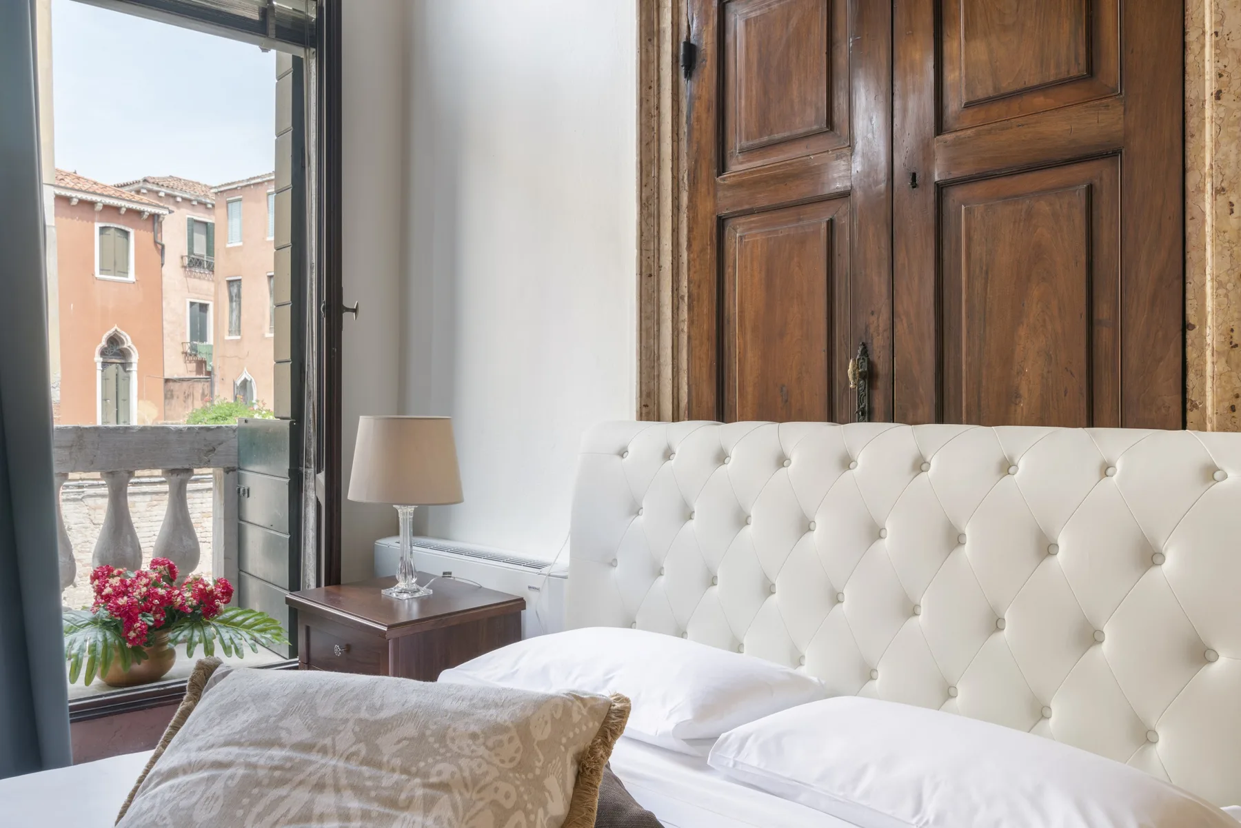 Un dormitorio con cabecero blanco capitoné, mesita de noche y ventana con vistas a edificios cercanos.