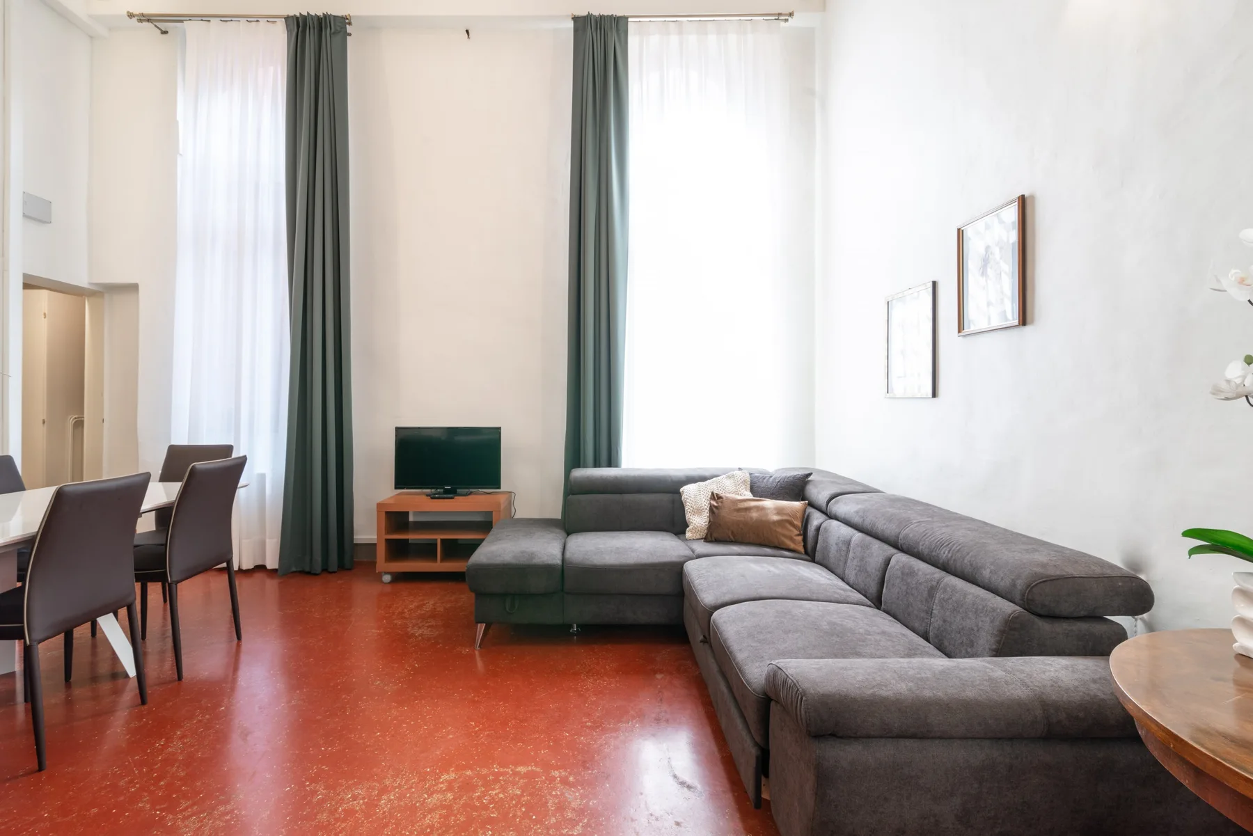 Una sala de estar con un sofá seccional gris y un televisor junto a ventanas altas.