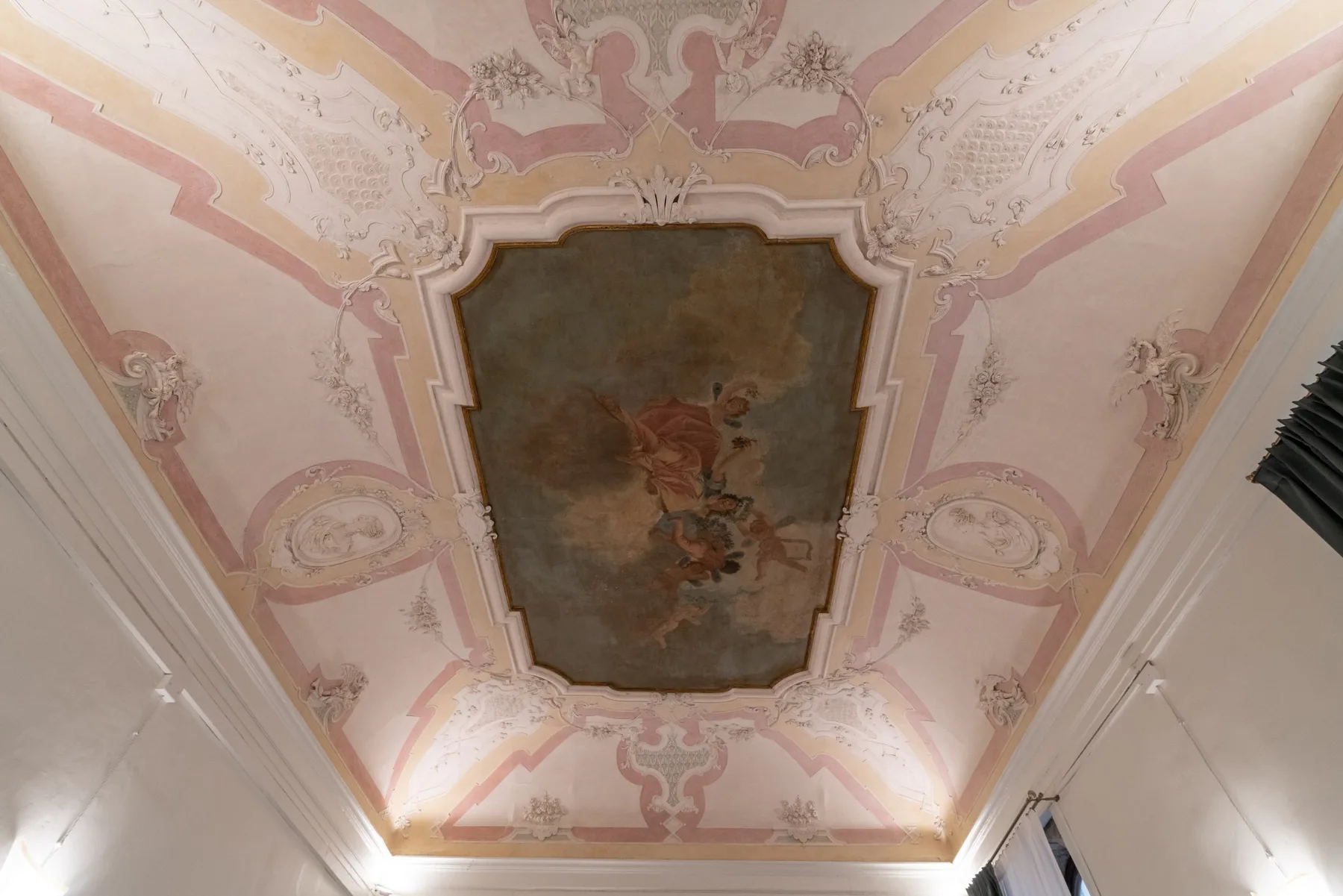 Un techo pintado y ornamentado presenta un mural central rodeado de detalles decorativos rosados.