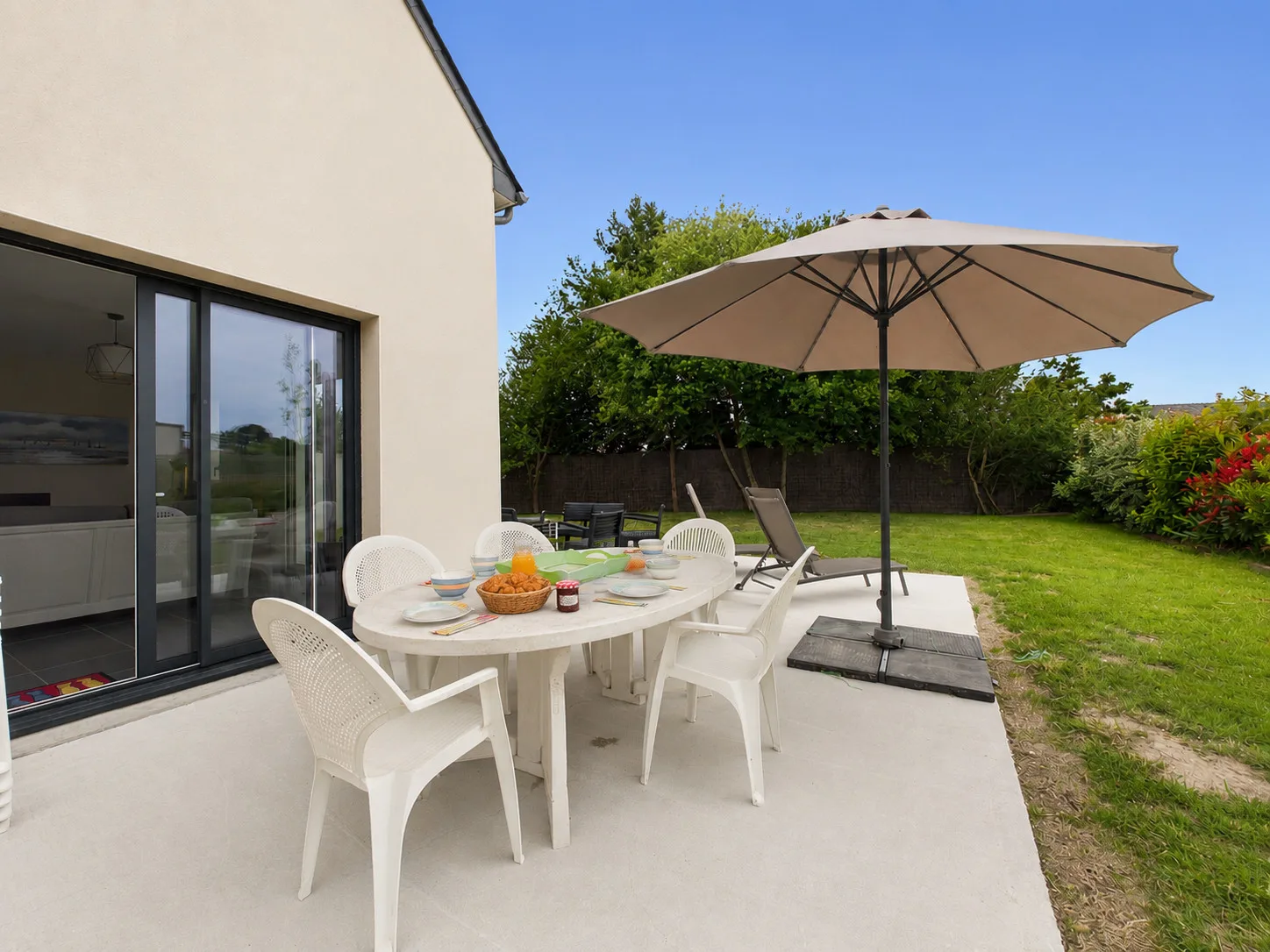 A patio dining area with white seating and a large umbrella beside a green lawn.