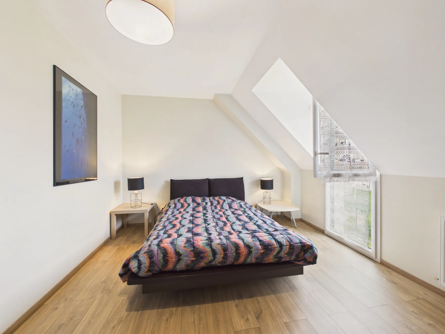 A bright bedroom with a bed beneath a sloped ceiling and a large window.