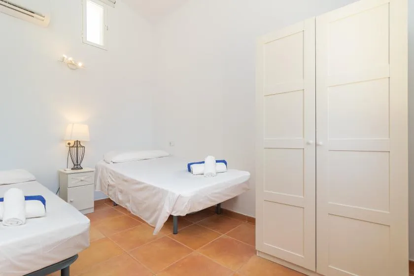 A bright bedroom with two beds, white linens, and terracotta-colored floor tiles.