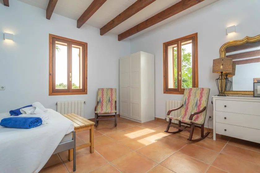 A bright bedroom with a bed, seating, and windows beneath exposed ceiling beams.