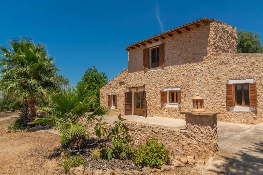 A stone house with wooden-framed windows stands among palm trees and garden plants.