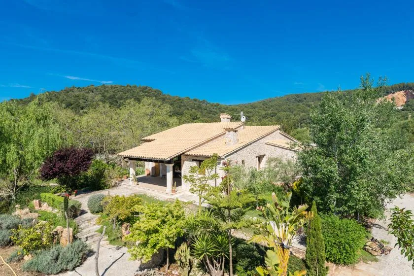 Una casa de tonos claros está rodeada de jardines, con colinas arboladas al fondo.