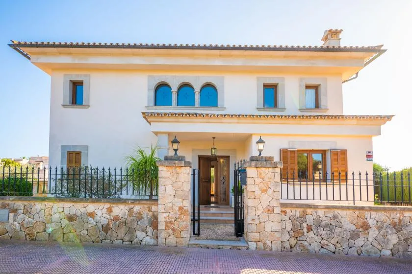 A white two-story house with arched windows, a gated entrance, and a stone wall.