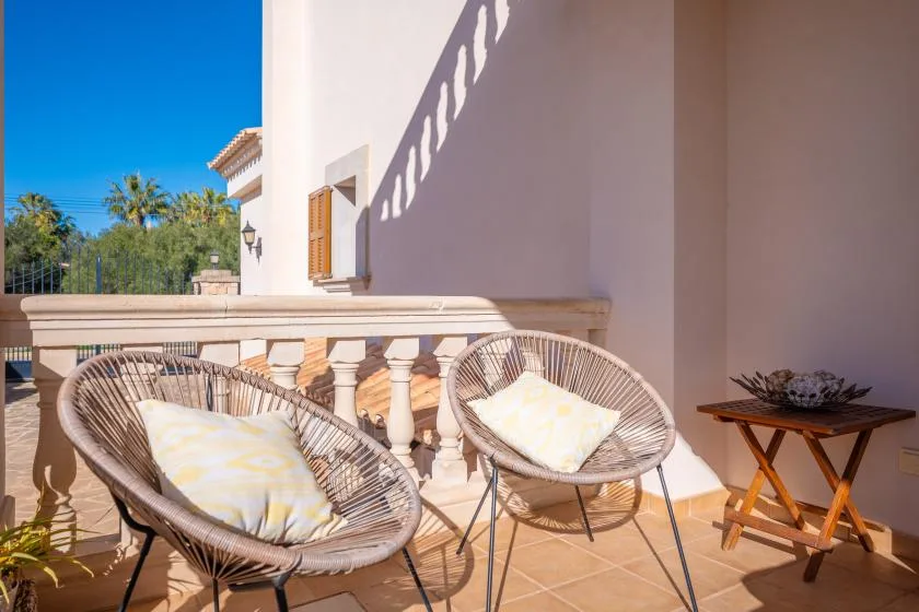 A sunlit balcony has two woven seats, a small table, and views of greenery.