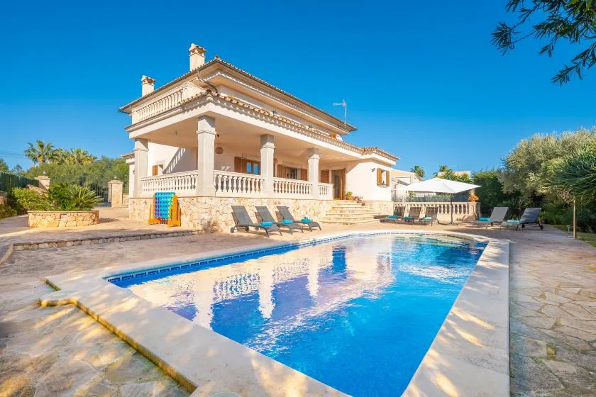 A swimming pool sits in front of a large house with outdoor seating nearby.