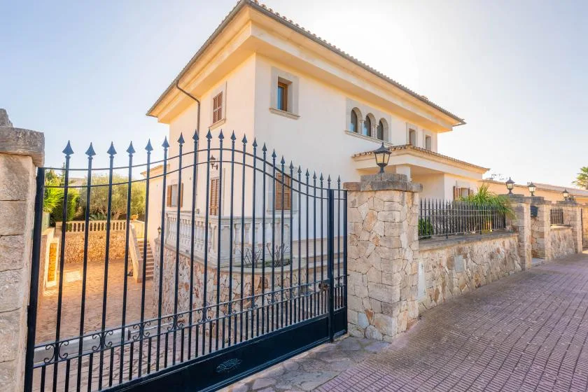A white two-story house stands behind a stone wall and black metal gate.