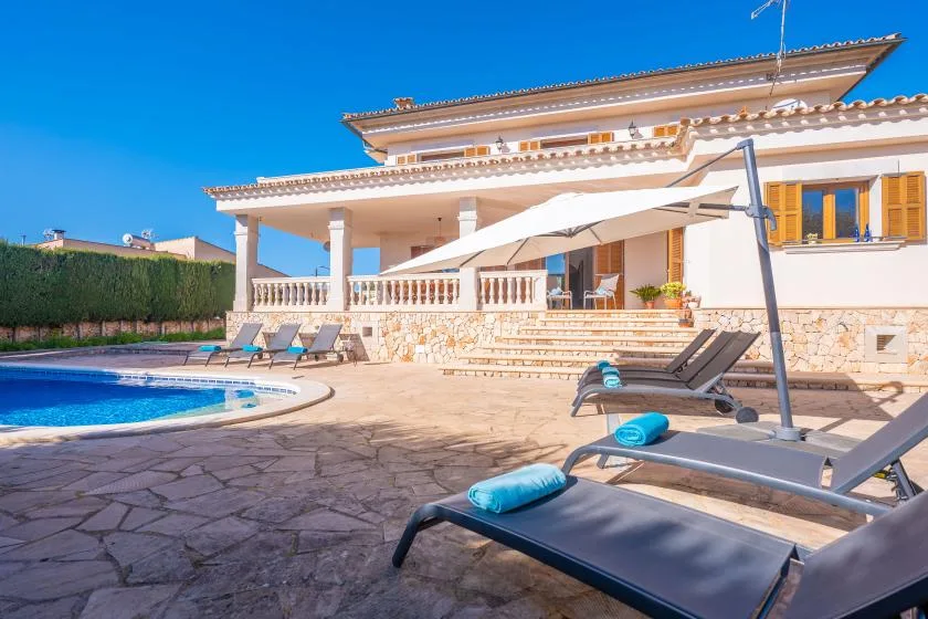 A swimming pool and sun loungers sit beside a house on a paved patio.