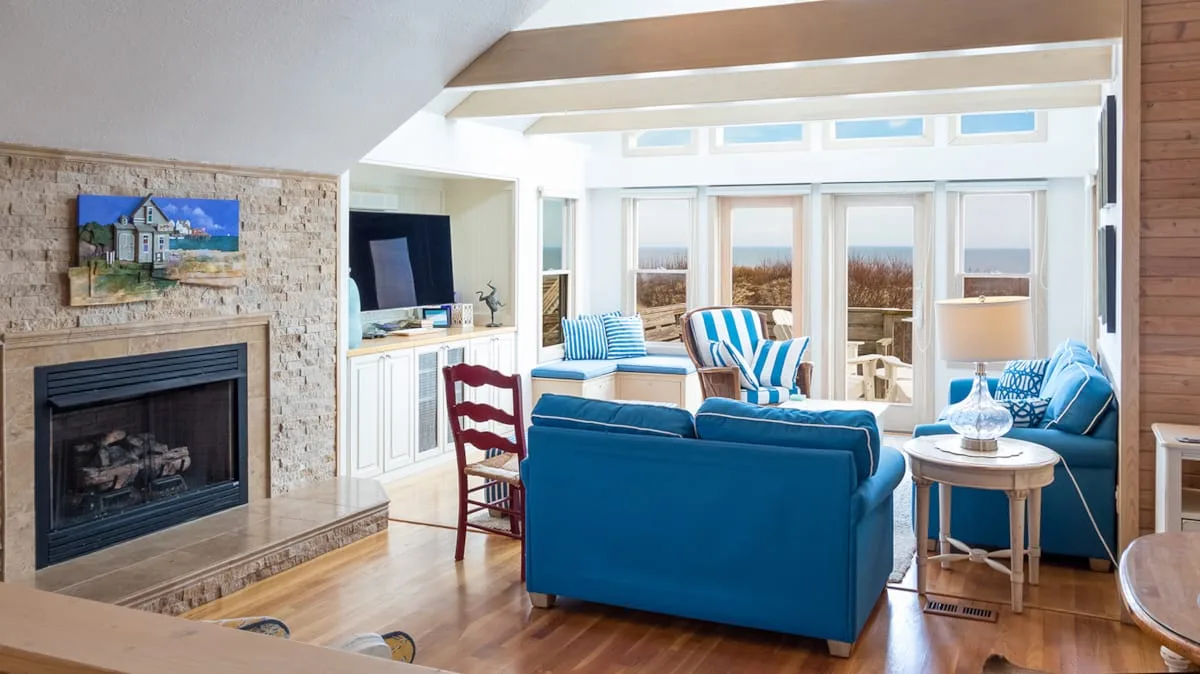 Una sala de estar luminosa con asientos azules, una chimenea y grandes ventanas.