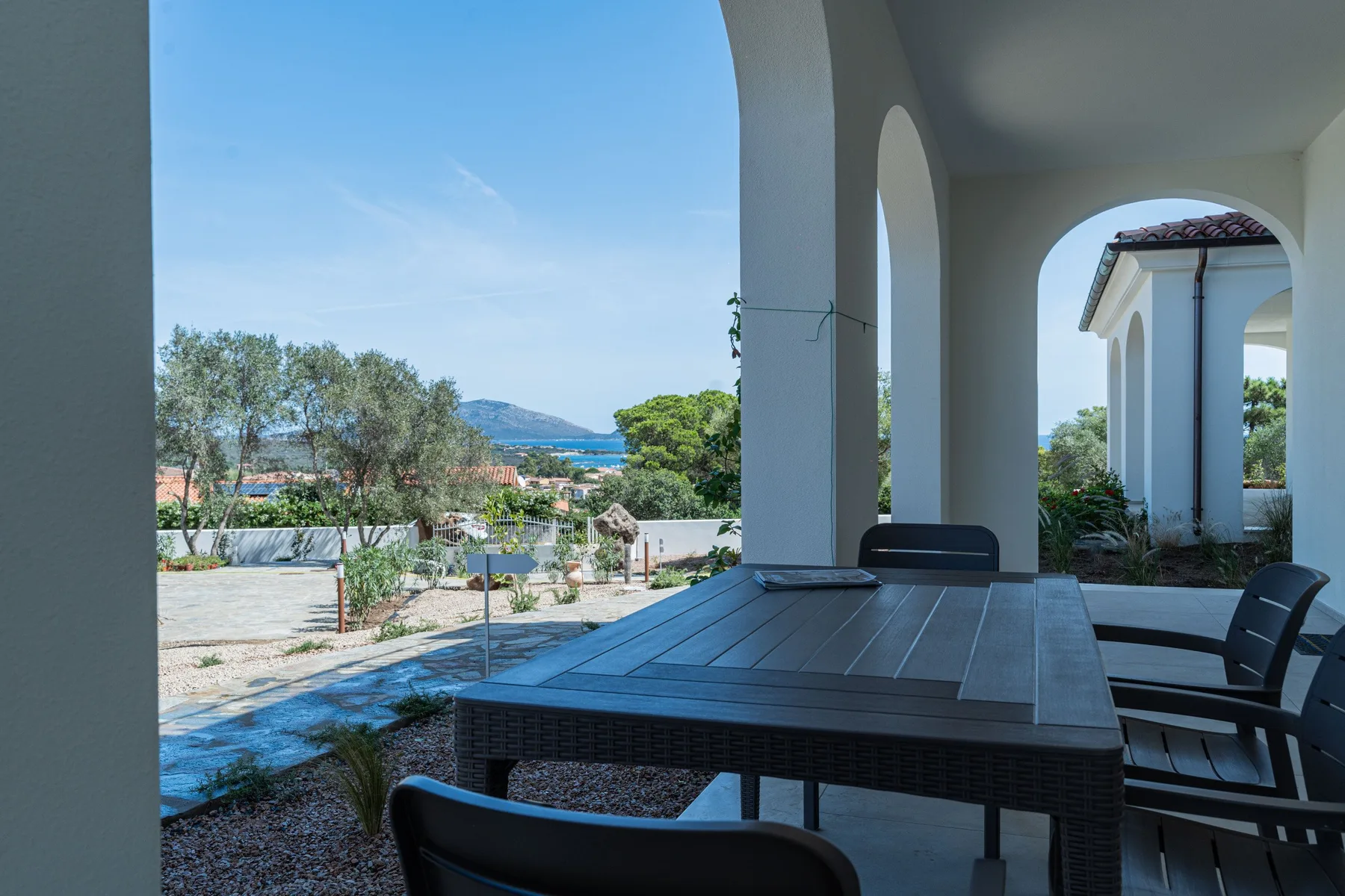 Una zona de comedor exterior a la sombra da a un jardín y al mar a lo lejos.