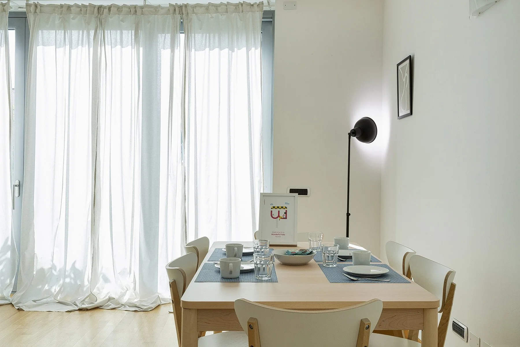 A dining area with a table set beside tall windows covered by sheer curtains.
