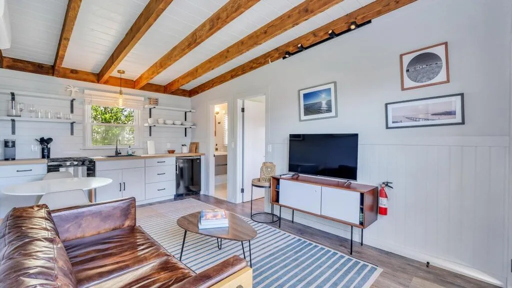 A bright living room with a television and an open kitchen beneath exposed wooden beams.