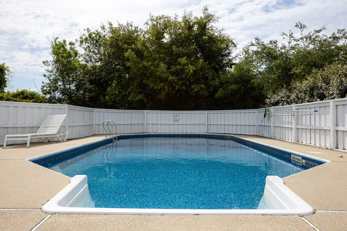 Una piscina al aire libre está junto a una cerca blanca y asientos de descanso.