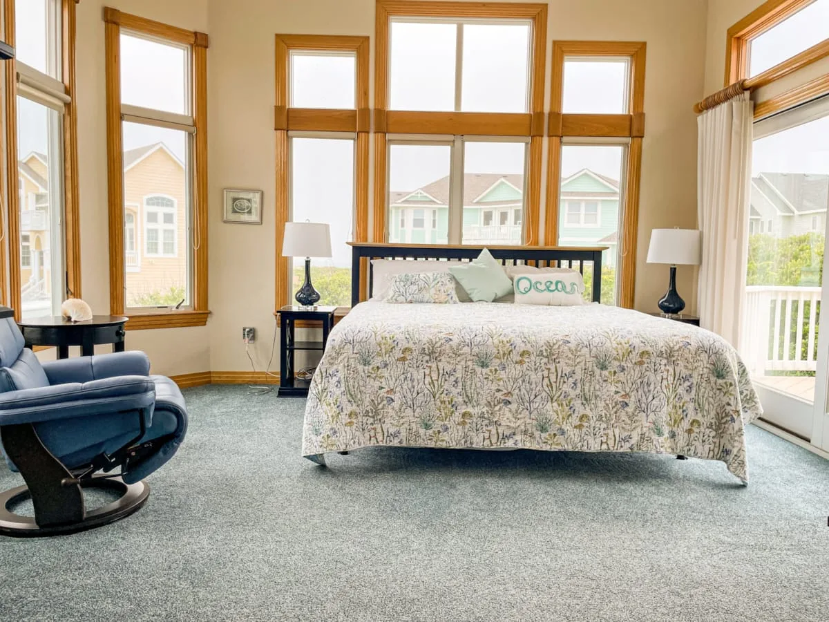 Un dormitorio luminoso tiene una cama junto a grandes ventanas y un sillón azul.
