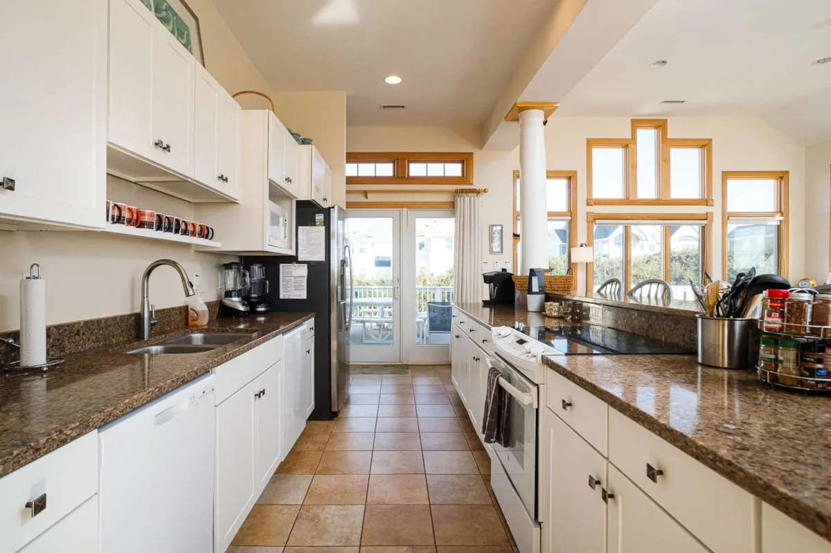 Una cocina luminosa con encimeras largas, gabinetes blancos y ventanas.
