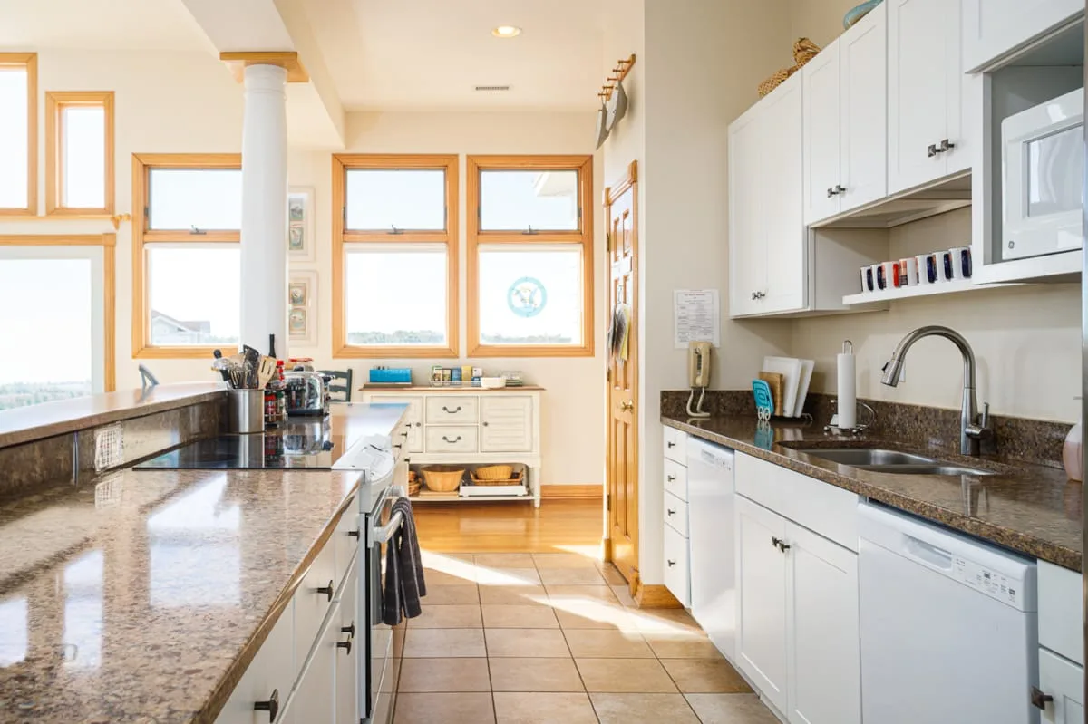 Una cocina luminosa con gabinetes blancos, encimeras oscuras y ventanas grandes.
