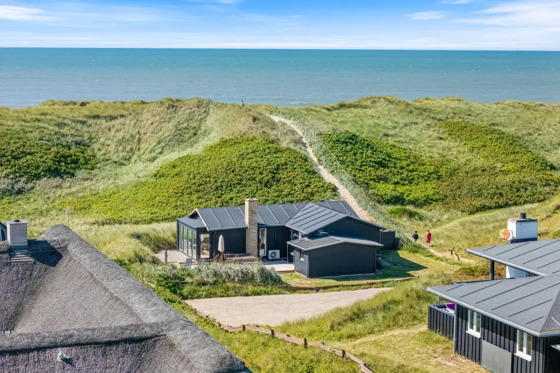 Vista aérea de una casa costera entre dunas cubiertas de hierba junto al mar.