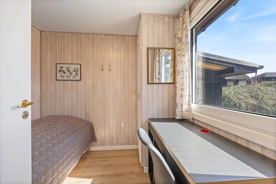 Un dormitorio con una cama, un escritorio bajo una ventana grande y paredes revestidas de madera.