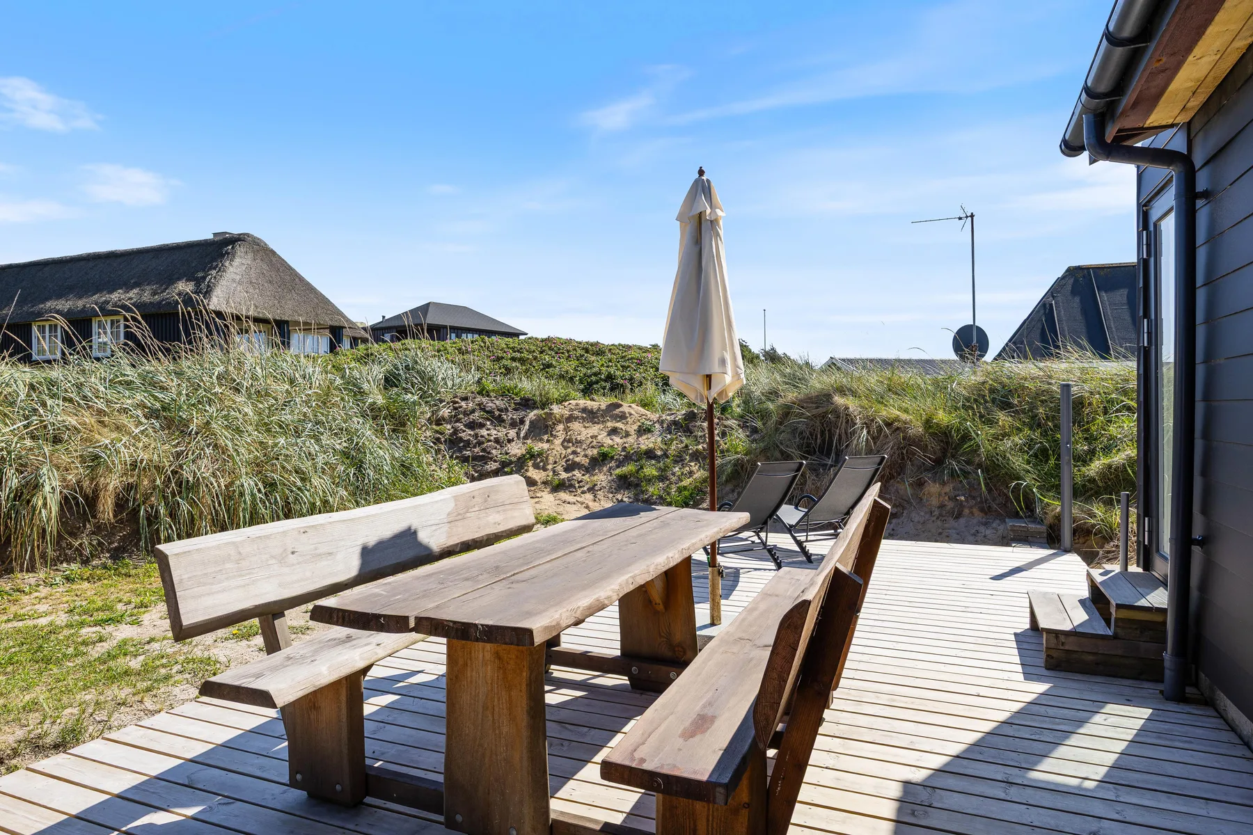 Una terraza de madera tiene una mesa de picnic, bancos y una sombrilla cerrada.