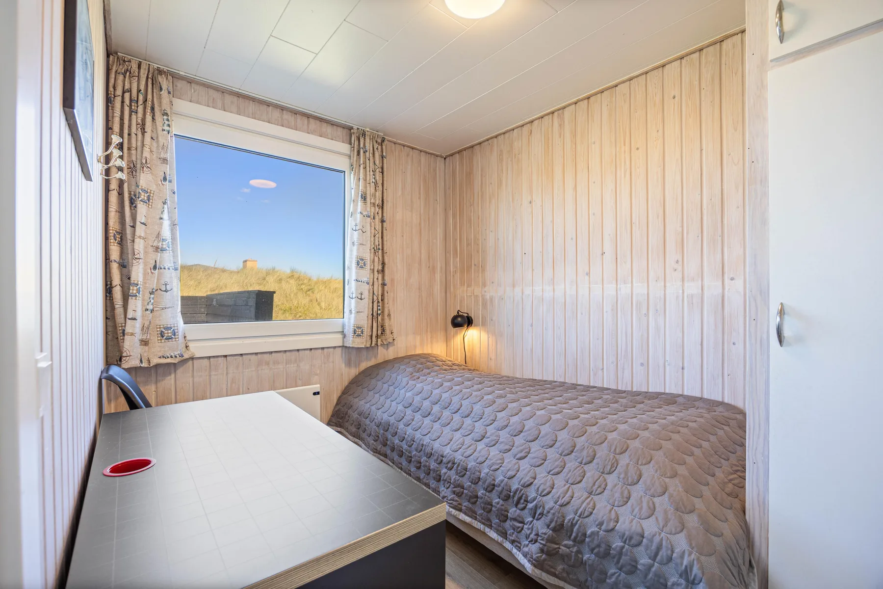 Un dormitorio pequeño con cama, escritorio y una ventana con vistas a dunas cubiertas de hierba.