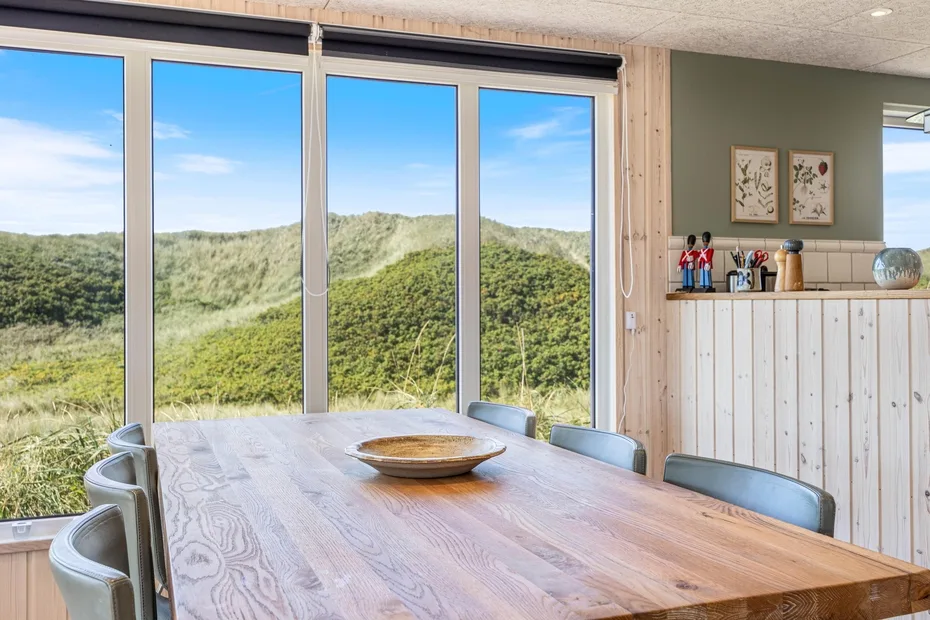 Comedor con una mesa de madera y grandes ventanas con vistas a colinas verdes.