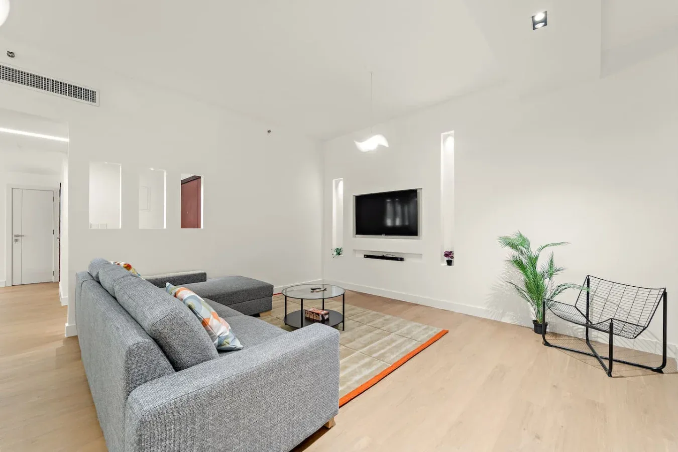 Una sala de estar luminosa con asientos grises, un televisor en la pared y una planta en maceta.