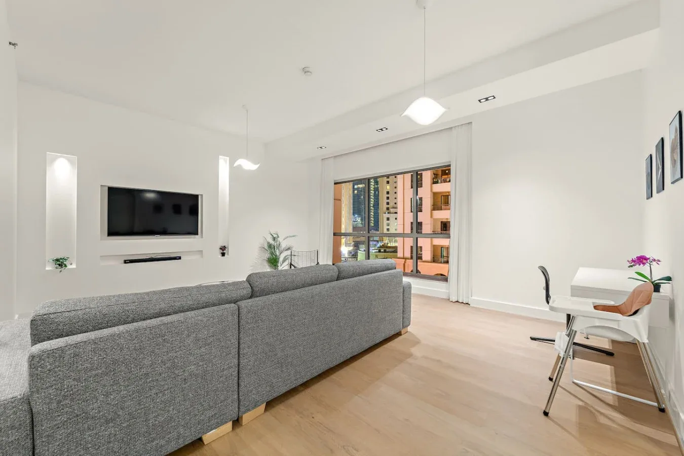 Una sala luminosa con asientos grises, un televisor en la pared y ventanas grandes.