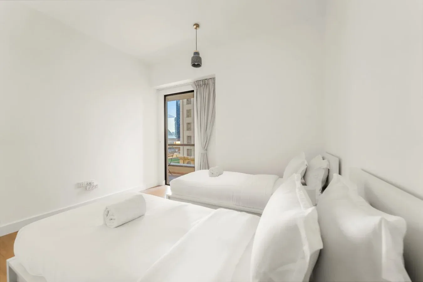 Un dormitorio luminoso con ropa de cama blanca y una ventana con vistas a edificios cercanos.