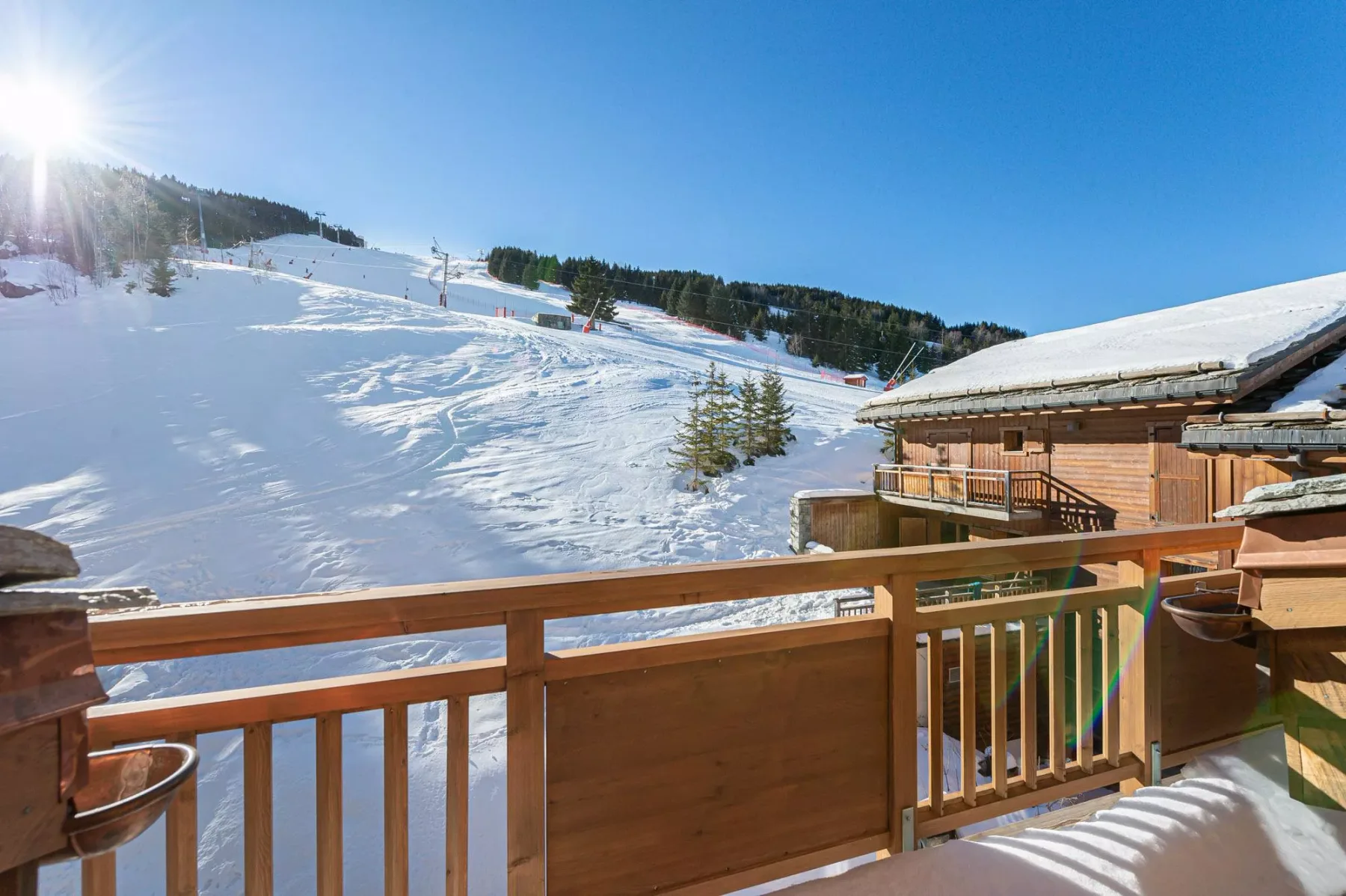 Una terraza de madera da a una pista de esquí nevada y un refugio bajo un cielo azul despejado.