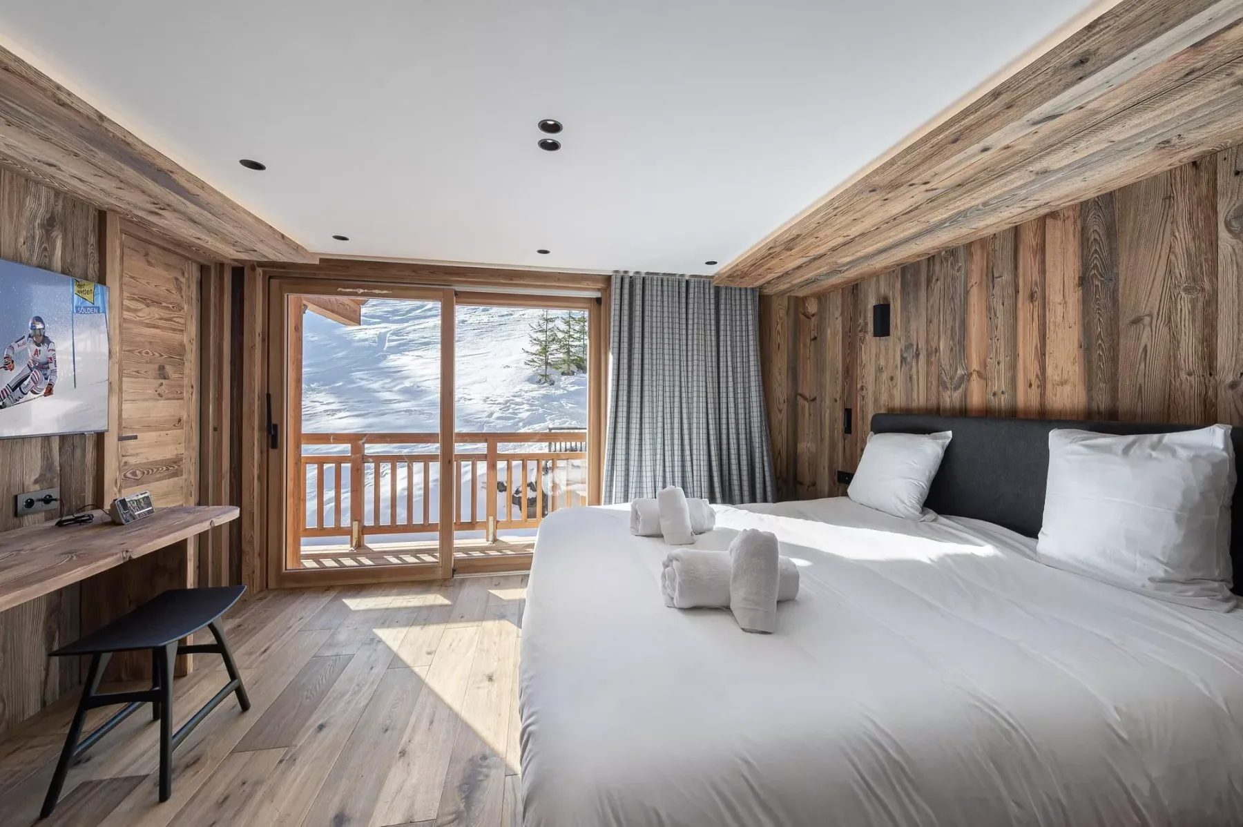 Un dormitorio con una cama grande, paredes revestidas de madera y una ventana al balcón con vistas a montañas nevadas.