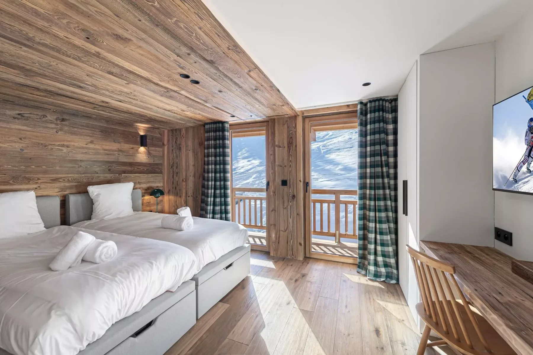 Un dormitorio con camas blancas, paredes revestidas de madera y ventanas con vistas a montañas nevadas.