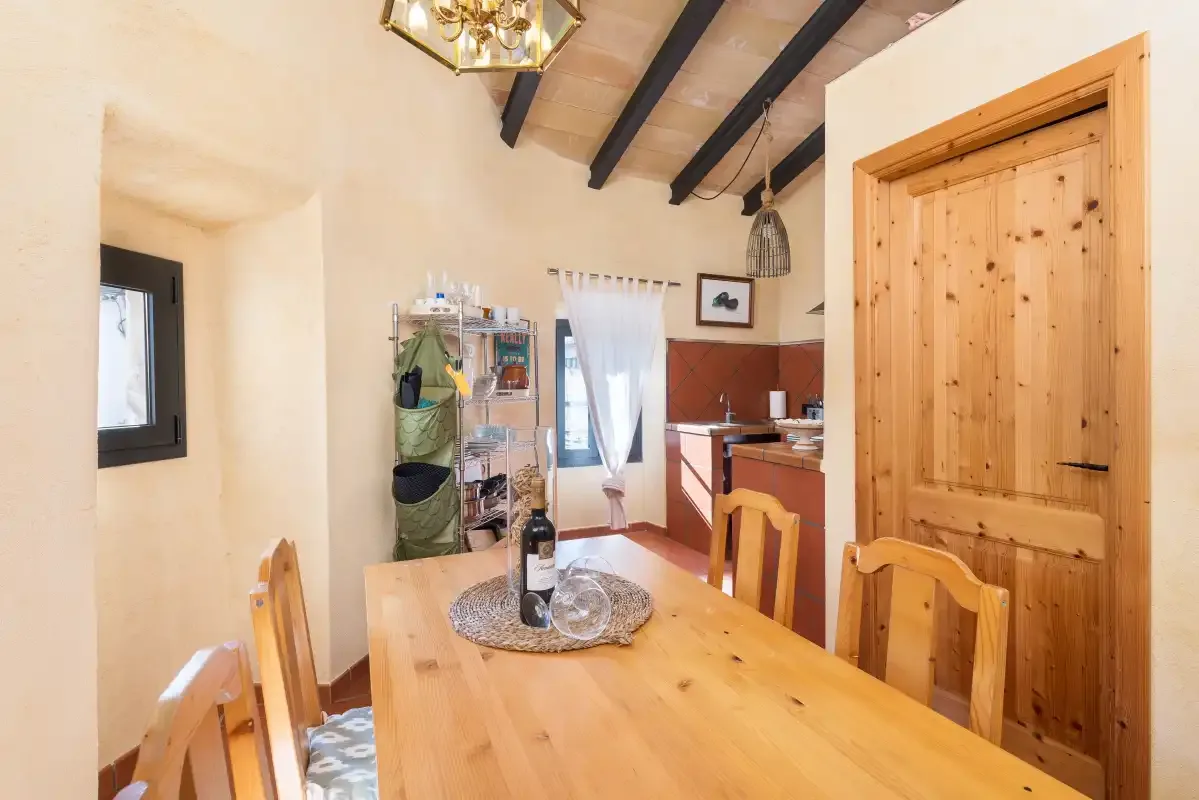A wooden dining table and chairs sit beside a kitchen in a bright room.
