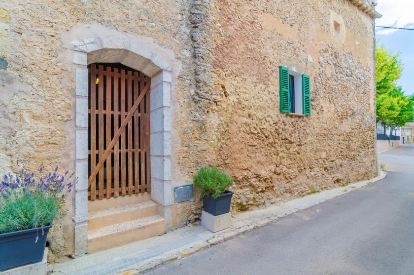 A stone building entrance opens onto a quiet paved street, with green shutters nearby.