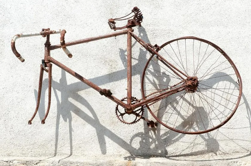 A rusty bicycle frame with a large rear wheel rests against a light wall.