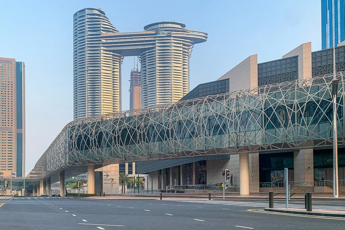 A modern city streetscape features a long glass-covered walkway and high-rise buildings.